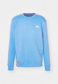 Sudadera de cuello redondo en azul claro con mangas largas, pequeño logo blanco de Alpha Industries en el pecho y etiqueta de tela roja cerca del dobladillo.