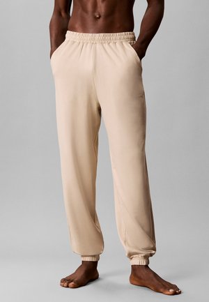 Personne torse nu portant un pantalon beige avec taille et chevilles élastiques, les mains dans les poches, debout pieds nus sur une surface grise.