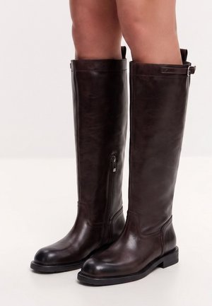 Dark brown knee-high leather boots with side zipper and buckle detail worn on bare legs against a plain light background.