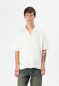 Kortærmet skjorte med knaplukning i struktureret off-white stof. Har en spids krave og en afslappet pasform. Parret med grønne denimjeans.