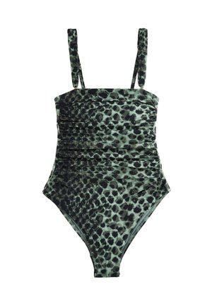 Woman in green patterned one-shoulder swimsuit and sunglasses stands by a pool with an ocean view in the background.