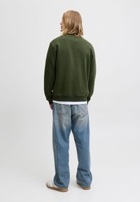 Blond person som står med ryggen mot kameran och bär en olivgrön sweatshirt, lösa urtvättade blå jeans, vita skor och en vit tröja under.