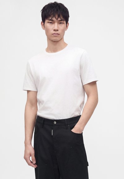 Jeune homme portant un t-shirt blanc uni à manches courtes et un pantalon noir, une main dans la poche, debout devant un fond blanc uni.