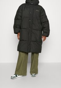 Zwarte pufferjas met een capuchon, drukknoopsluiting, gequilte structuur en een logo op de borst. Gecombineerd met olijfgroene wijde broek en sneakers.