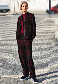 Schwarze und rote karierte Bomberjacke über einem maroone Pullover, kombiniert mit passenden karierten Hosen und schwarzen Sneakers mit weißen Sohlen.