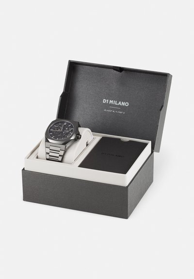 Schwarze Uhr mit geometrischem Design an einem silbernen Edelstahlarmband, präsentiert in einer dunkelgrauen Box mit einem weißen Kissen und Markenlogo.