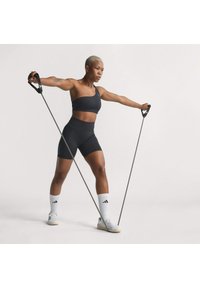 Sportiva nera e pantaloni corti, fasce di resistenza elastiche in mano, in piedi su una gamba. Sfondo minimalista, colori neutri, calzature sportive.