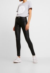 Pantalones skinny de piel sintética negra con una textura suave, combinados con una camiseta gris claro con un gráfico circular y zapatillas blancas.