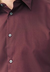 Bordeaux knap-up skjorte med struktureret stof, der har en klassisk krave og kontrasterende blå knapper. Glat finish med en subtil glans.
