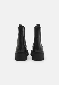 Botas de tobillo de cuero negro con una textura suave, paneles laterales elásticos, lengüetas y suelas de goma gruesas. La vista trasera enfatiza la altura y el diseño.