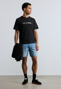 Ung mand iført sort Calvin Klein t-shirt, slidte denimshorts, sorte sneakers og holder en sort sweater, stående mod en ensfarvet lys væg.