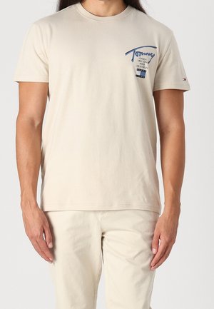 Hombre que lleva una camiseta beige de manga corta y pantalones beige a juego, con texto y pequeño logo en el pecho izquierdo y la manga.
