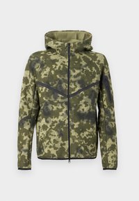 Kapuce jaka ar zaļo kamuflāžas rakstu. Izgatavota no teksturēta auduma ar rāvējslēdzēja aizdari un melnām akcentu šuvēm. Iezīmēta ar pieguļošu dizainu.