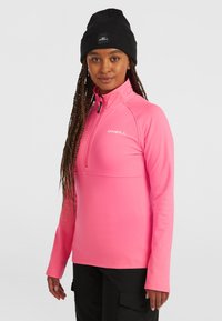 Sudadera de forro polar con media cremallera en un rosa brillante, hecha de material ligero. Presenta un cuello alto y un detalle de logo en el pecho. Combinada con un gorro negro.