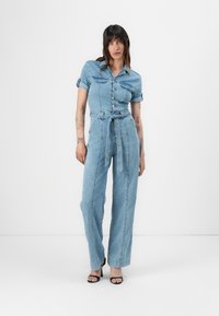 Denim kombinezon v svetlo modri barvi z oprijetim zgornjim delom s kragnico, zapenjanjem na gumb in širokimi hlačnicami. Vključuje pas z obrobo in zavihane rokave.