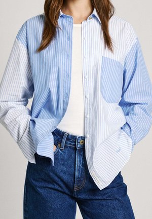 Kobieta ubrana w niebiesko-białą pionowo prążkowaną koszulę na guziki, założoną na biały top, z rękami włożonymi do kieszeni wysokich niebieskich dżinsów.