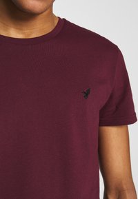Burgunderfarbendes Rundhals-T-Shirt aus weicher Baumwolle mit kurzen Ärmeln, versehen mit einem kleinen schwarzen gestickten Vogel-Logo auf der linken Brust.