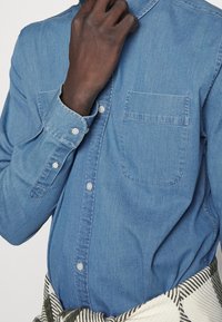 Camisa de denim azul claro con cuello abotonado, bolsillo en el pecho y mangas largas, con un ajuste clásico y una textura sutil.