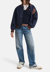 Blouson aviateur rayé bleu marine avec des accents orange, pull en maille torsadée bleu marine, jean bleu clair, et baskets blanches avec des bandes noires.