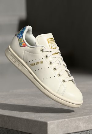 Hvid Adidas Stan Smith-sneakers med guldtekst, perforeringer på siden og et farverigt blomstermønster på hælen, svævende over en stenovertflade.