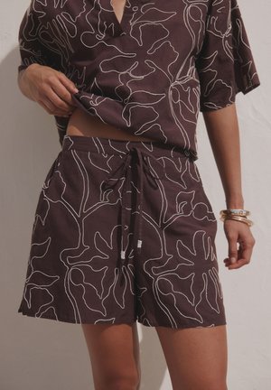 Person iført matchende mørkelilla shorts og kortærmet top med hvide abstrakte linjemønstre, løfter let trøjen, armbånd om håndleddet.