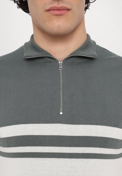 Ribbad stickad tröja med dragkedja i mörkgrön med vita horisontella ränder. Har en ståkrage och ribbad textur. Detalj i silvermetall på dragkedjan synlig.