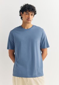 Hele sinine lühikeste varrukatega T-särk, valmistatud siledast kangast, millel on ümmargune kaelus ja lahtine lõige. Lihtne disain ilma nähtavate mustrite ega aksentideta.