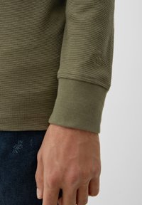 Maglietta a maniche lunghe color verde oliva con un design a tessuto waffle testurizzato, caratterizzata da un polsino aderente e un sottile logo sulla manica.