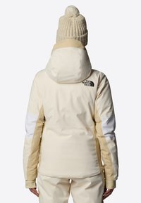 Giacca da neve color crema con cappuccio, con pannelli beige e bianchi. Texture morbida con il logo The North Face sul retro, polsini a coste.