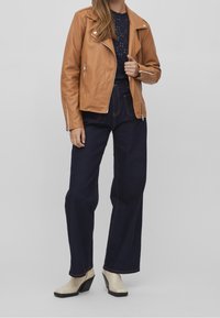 Brązowa skórzana kurtka motocyklowa z srebrnymi detalami, granatowa bluzka, szerokie ciemnoniebieskie dżinsy oraz beżowe botki na obcasie.