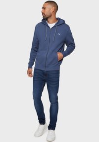 Blaue Zip-Hoodie aus Baumwollmix, mit Kängurutasche, Kordelzügen und Logodetail auf der Brust. Getragen mit Jeans und Sneakern.