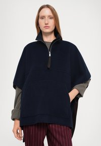 Una giovane donna con capelli rossi lisci indossa un poncho blu navy con mezzo zip, maniche lunghe grigie e pantaloni bordeaux a righe, in piedi contro uno sfondo bianco.