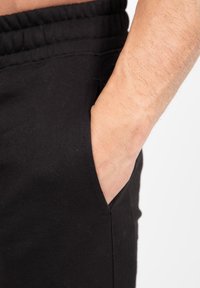 Juodos medvilnės sportinės kelnės su elastinga juosmens juosta ir šoniniu kišenėle, rodančia ranką, ilsinčiąsi kišenėje, atskleidžiančia lygią tekstūrą.