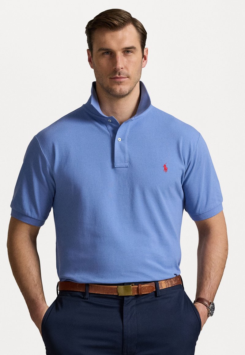 Svijetloplava polo majica s kratkim ovratnikom, patent zatvaračem na dva gumba i crvenim logotipom na prsima, uparena s tamnim hlačama i smešnim remenom.