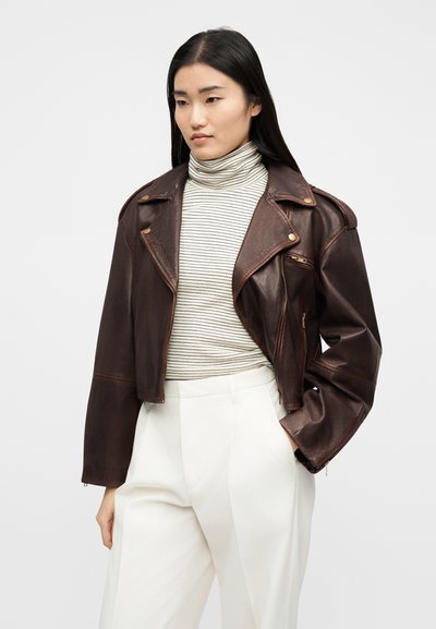 Γυναίκα με cropped σκούρο καφέ δερμάτινο μπουφάν πάνω από ένα ανοιχτόχρωμο ριγέ ζιβάγκο και λευκό παντελόνι ψηλόμεσο με φαρδιά πόδια, στέκεται με το ένα χέρι στην τσέπη.