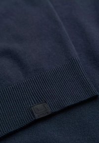 Pull en maille bleu marine avec ourlet côtelé, texture douce et petite étiquette noire avec logo dans un coin en bas.