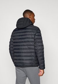 Juodas pūkinis švarkas su siuvinėta tekstūra ir gobtuvu. Lygi tekstūra, elastingos rankovės ir užtrauktukas priekyje. Dėvimas ant pilkų sportinių kelnių.