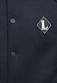 Πλεκτό μπουφάν σε ναυτικό μπλε με κουμπιά τύπου σναπ και υφασμάτινο διακοσμητικό σε σχήμα ρόμβου που έχει λευκό 'L' και τη λέξη 'LACOSTE'.