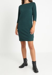 Vestido acolchado en tono verde azulado con mangas tres cuartos, cuello redondo y diseño ajustado, combinado con botas negras hasta el tobillo. Textura suave con patrón en relieve.
