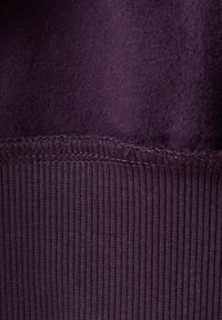 Nahaufnahme eines dunkelvioletten Stoffs, der den Übergang von einer weichen Fleece-Oberfläche zu einem gerippten Strick zeigt, mit sichtbaren Nähten.