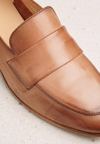Mocasín de piel marrón con una correa ancha y plana en el empeine; textura suave; forma redondeada; detalles de costura mínimos a lo largo de los bordes.