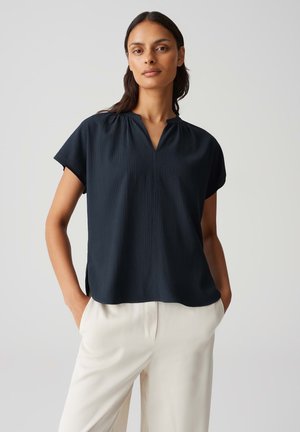 Blusa blu navy a maniche corte con scollatura a V, caratterizzata da un tessuto testurizzato e lievi pieghe sulle spalle. Indossata con pantaloni di colore chiaro.