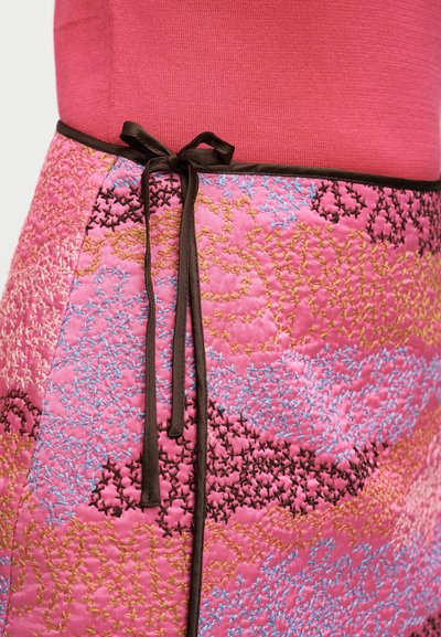 Ροζ υφασμάτινο κουβερλί με πολύχρωμα κεντημένα μοτίβα σταυροβελονιάς, δεμένο με μαύρο φιόγκο κορδέλας στη μέση πάνω από ροζ τοπ.