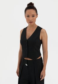 Black pinstriped vest with a V-neck, five buttons, and cropped design. Worn with matching trousers, featuring an angled cut and subtle brand tag.
