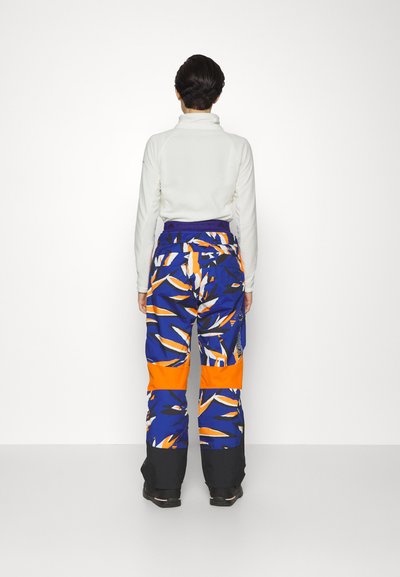 Kleurige skibroeken in blauw, oranje en wit met een bladpatroon, met een hoge taille en zwarte boorden bij de enkels.