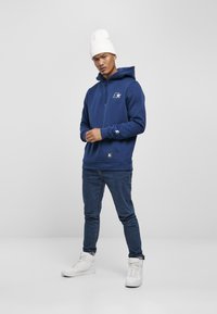 Hoodie blu navy in morbido tessuto con cordino, logo sul petto, abbinato a jeans slim fit e sneakers alte bianche.