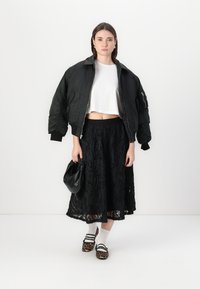 Veste bomber noire avec des poignets côtelés, t-shirt blanc court, jupe noire texturée, sac à main noir et chaussures à imprimé léopard avec des chaussettes blanches.