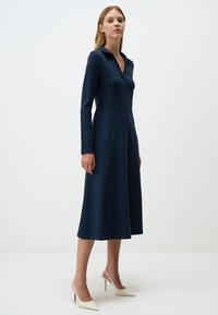 Robe bleu marine à manches longues avec col, corsage ajusté, jupe évasée, fabriquée à partir d'un tissu texturé. Complétée par des chaussures à talons hauts blanches.