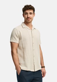 Kortærmet beige skjorte med knapper, lavet af tekstureret stof, med spredt krave og løs pasform, parret med mørkeblå bukser.
