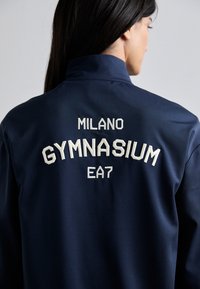 Navy blue sports jacket featuring embroidered text “MILANO GYMNASIUM EA7” on the back, made from smooth, lightweight fabric.